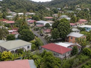 Aerial view of a hillside residential neighborhood with one and two-story homes, metal roofs, palms, and green hills in Saint Lucia