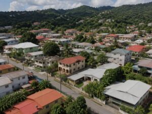 Aerial view of a residential neighborhood with one and two-story homes, metal and concrete roofs, trees, and green hills in Trinidad and Tobago