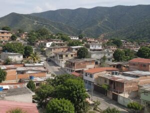 Aerial view of a present-day hillside residential neighborhood of brick and painted homes with metal and flat concrete roofs, mango trees and green mountains beyond in Venezuela