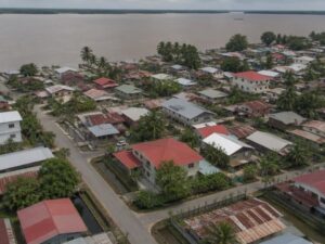 Aerial view of a residential neighborhood with wooden and concrete homes, metal roofs, palms and a broad brown river in Suriname