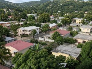 Aerial view of a hillside residential neighborhood with one and two-story homes, metal and concrete roofs, trees, and green hills in Jamaica